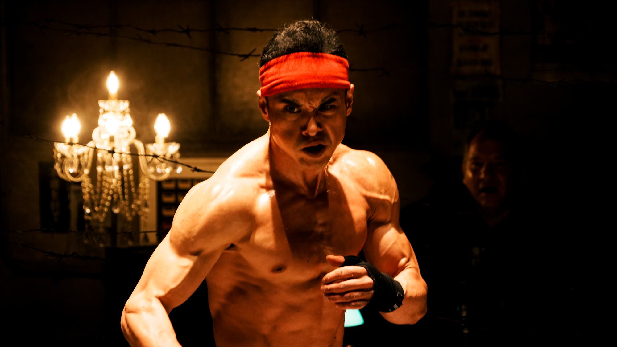 A shirtless fighter in a red headband under a chandelier